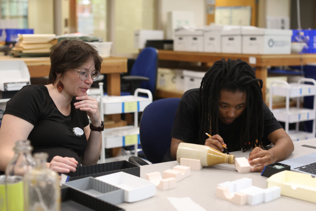 A professor sits with a student who is holding a pencil and working with artifacts and storage boxes.