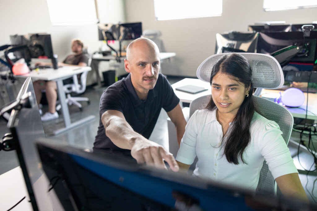 A student and a professor sit behind a computer, while the professor points something out on the screen.