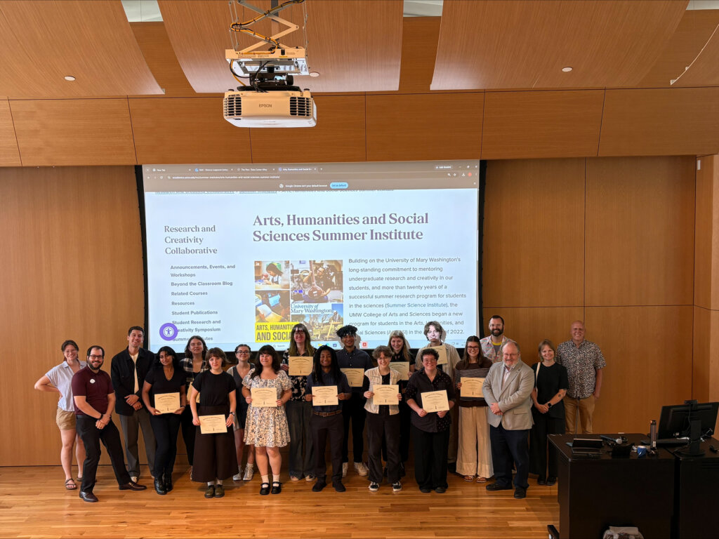 A group of students and faculty members stand in front of a screen projecting an image that represents UMW's Arts, Humanities and Social Sciences Summer Institute.