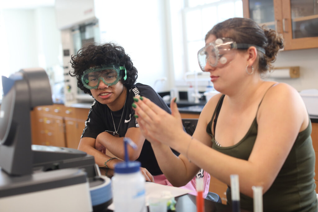 A girl and a boy wearing goggles sit next to each other inside a science lab