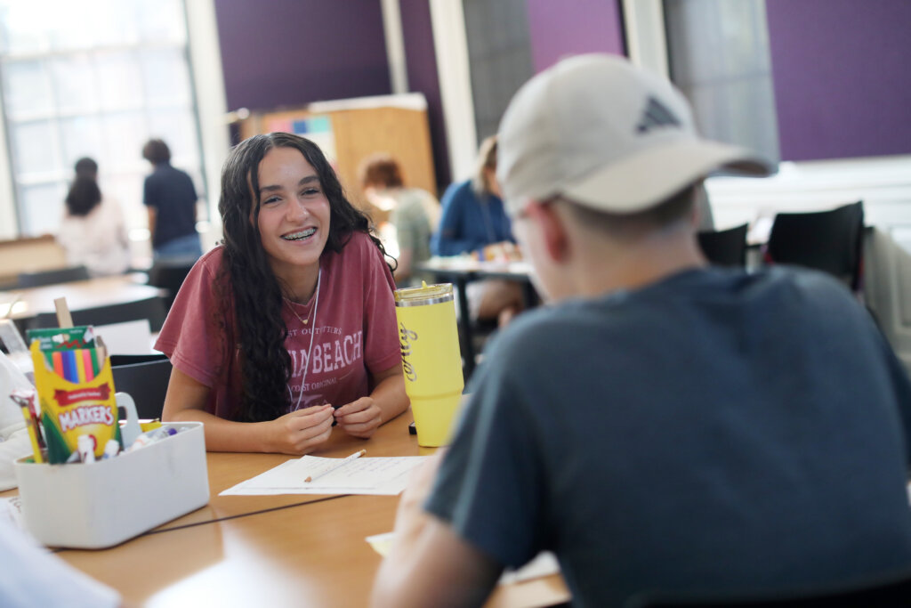 A girl in a college classroom with brightly colored magic markers smiles while talking to a guy