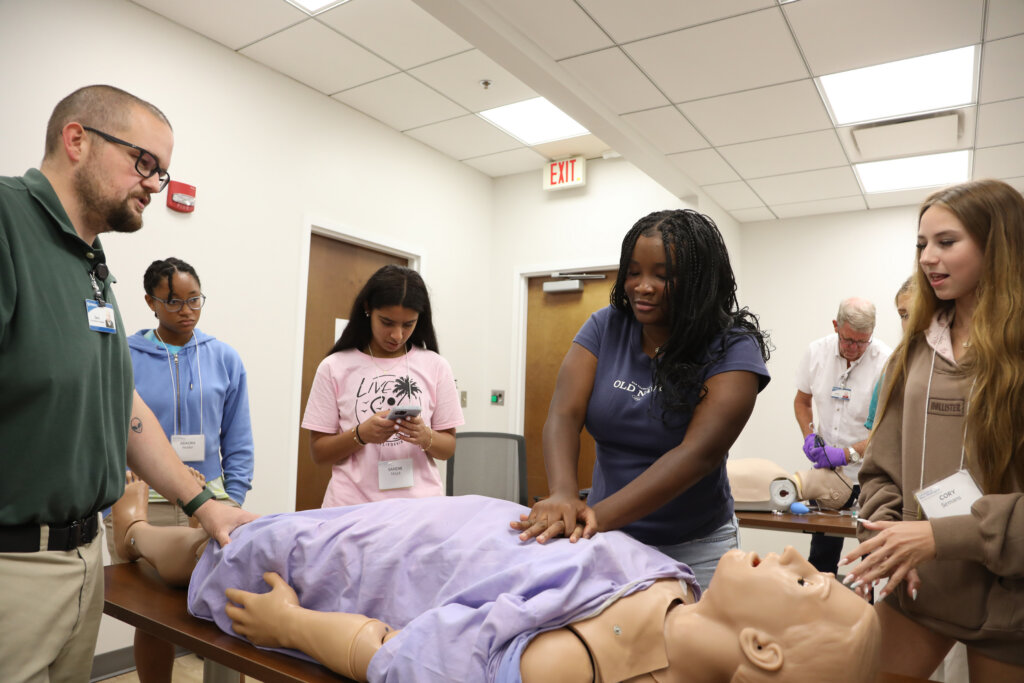 A student performs chest compressions on a fake patient while other students and healthcare professionals observe