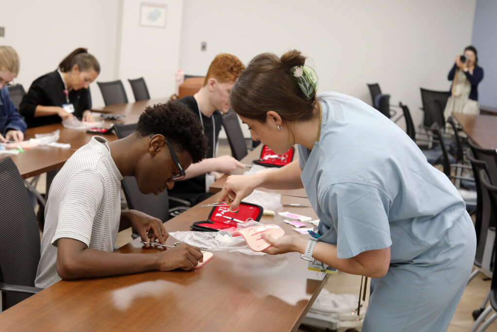 A woman in blue scrubs holds a surgical instrument and a pink suture-practice pad while helping a student learn to sew stitches