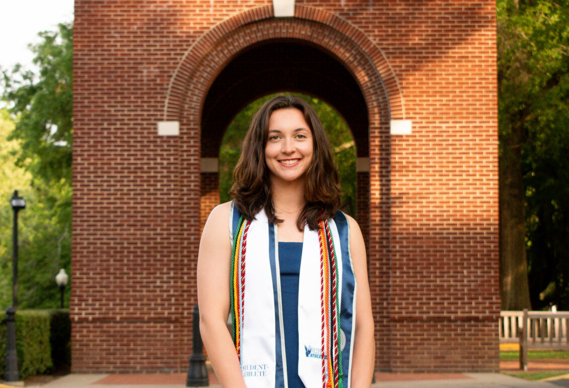 A young woman in college regalia stands in front of a brick structure