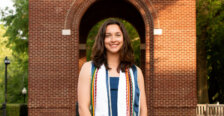 A young woman in college regalia stands in front of a brick structure