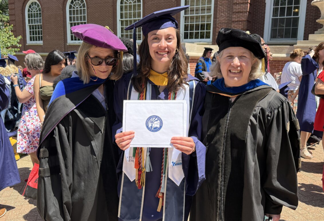 Three young women in college graduation regalia pose, while the one in the middle holds up her recently earned diploma