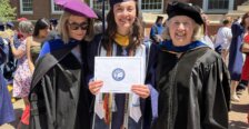 Three young women in college graduation regalia pose, while the one in the middle holds up her recently earned diploma