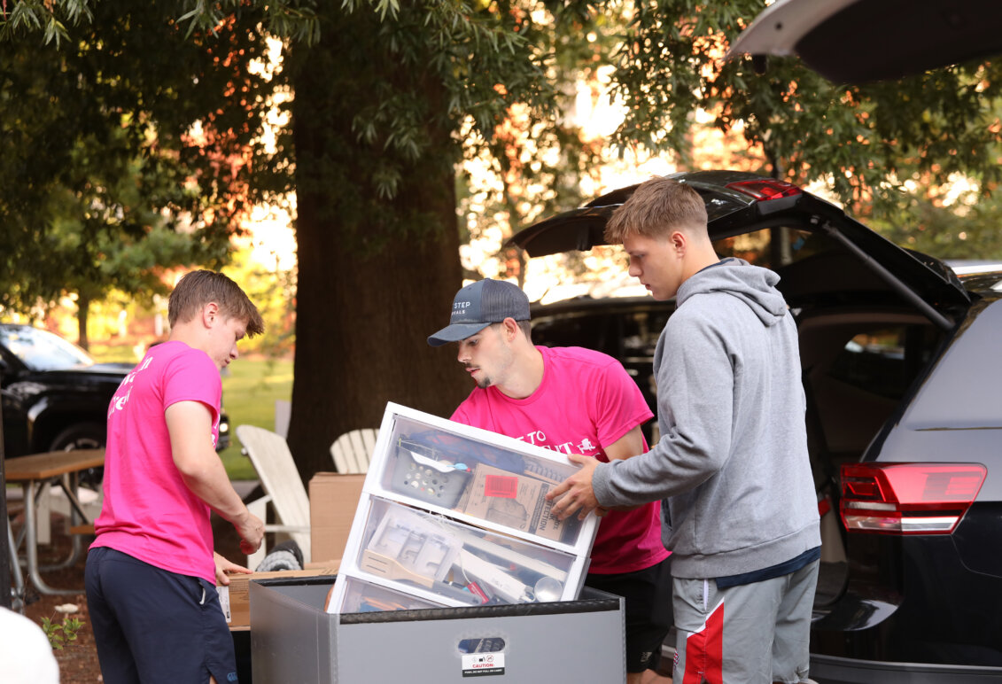 Two guys dressed in hot pink help another guy in a gray hoody load supplies to move into his residence hall room