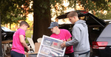 Two guys dressed in hot pink help another guy in a gray hoody load supplies to move into his residence hall room 
