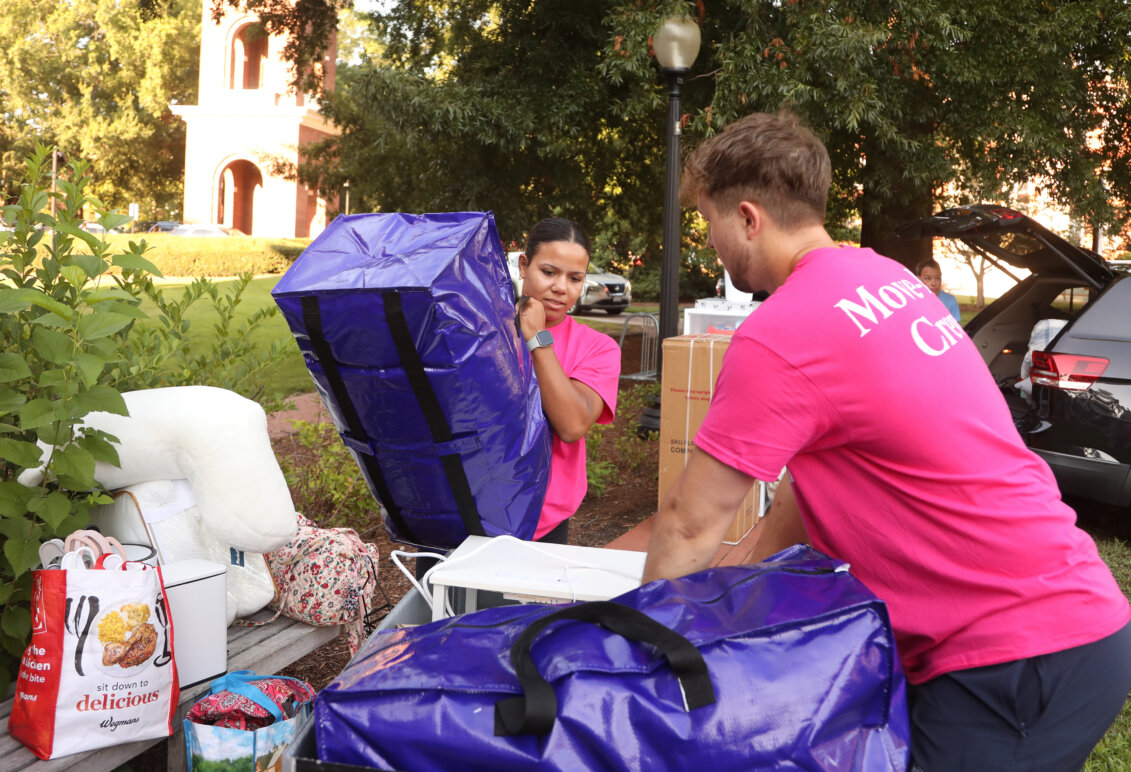 Students stand outside on a university campus wearing hot-pink T-shirts and holding big purple bags, while other bags surround them