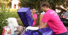 Students stand outside on a university campus wearing hot-pink T-shirts and holding big purple bags, while other bags surround them