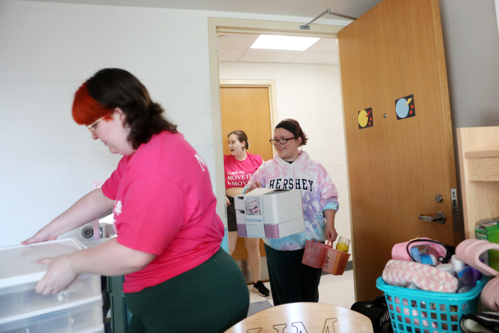 A girl puts a clear stack of drawers into a residence hall room, while another comes through the door carrying a couple of bathroom caddies