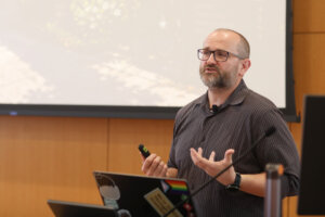 A man stands at a podium with a laptop in front of him and a screen behind him