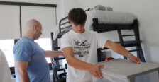 A young man puts together a clear multi-drawer unit, while an older man points to the bunkbed in the backrgound