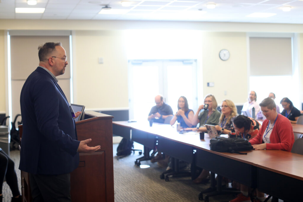 A man stands at the front of a room speaking to several people who are seated before him