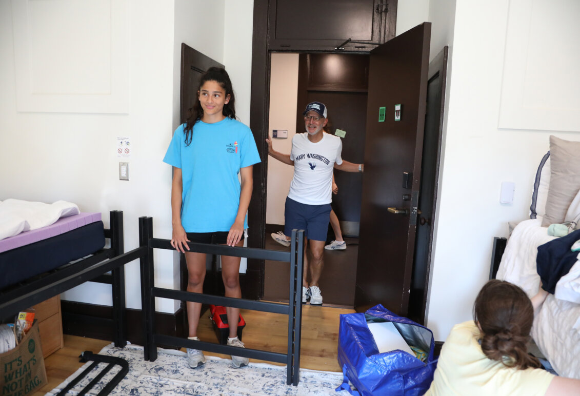 A man stands in the doorway to a room, while a young woman works on putting together a bed frame and another looks on