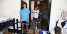 A man stands in the doorway to a room, while a young woman works on putting together a bed frame and another looks on