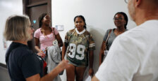 Five people - one man and four women - stand in a hallway talking