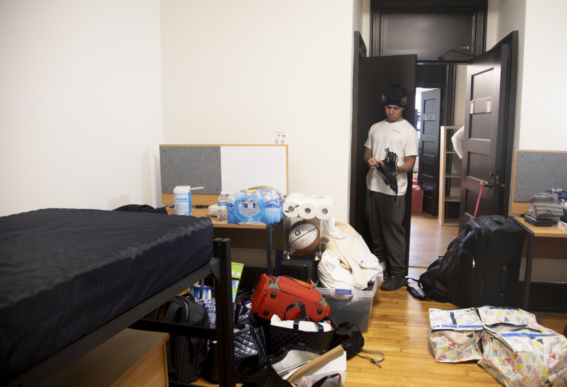 A young man stands outside a closet in a residence hall room with an array of items - paper towels, bottled waters, suitcases - in the foreground