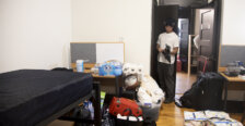 A young man stands outside a closet in a residence hall room with an array of items - paper towels, bottled waters, suitcases - in the foreground