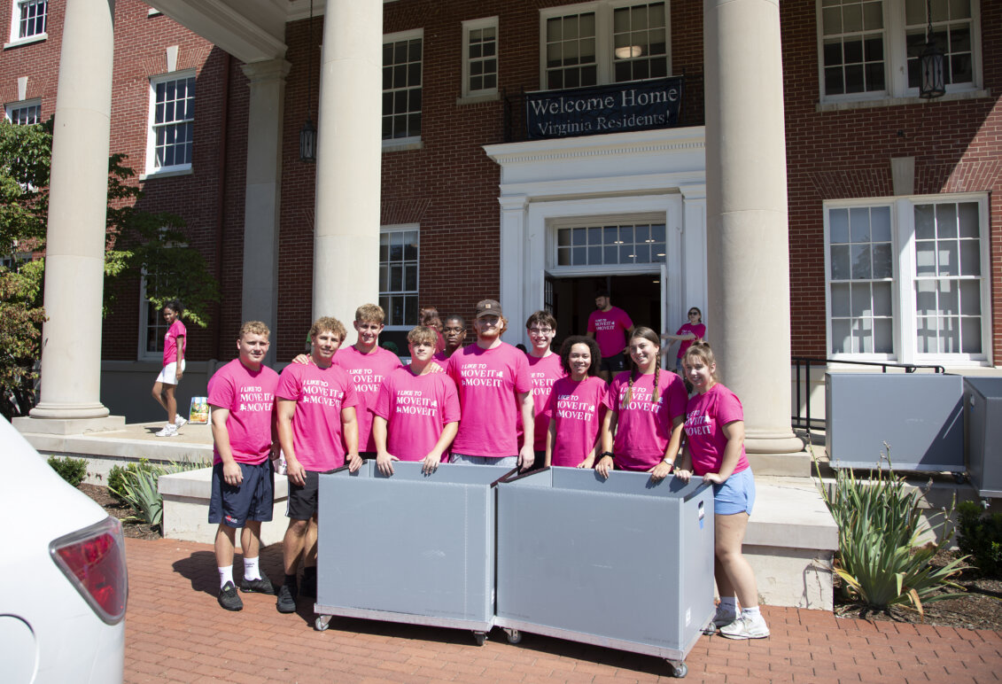 Several students dressed in hot pink T-shirts stand in front of a brick building with columns, with large gray rolling carts in front of them