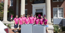 Several students dressed in hot pink T-shirts stand in front of a brick building with columns, with large gray rolling carts in front of them