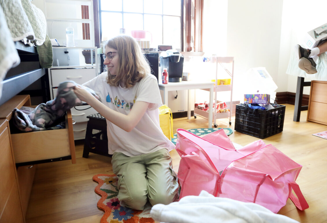A student sits on the floor of a residence hall, pulling something out of a drawer under the bed