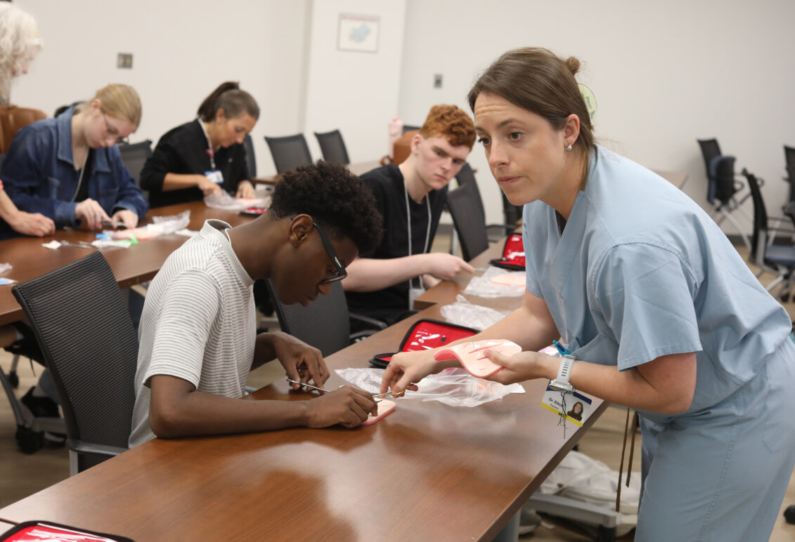 In a classroom a female doctor helps a student learn to suture while other students in the room also practice. Photo by K Pearlman Photography.