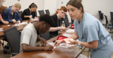 In a classroom a female doctor helps a student learn to suture while other students in the room also practice. Photo by K Pearlman Photography.