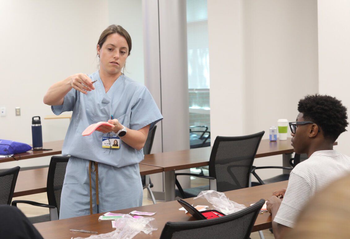 A female doctor in light blue scrubs in a classroom demonstrates suturing to high school students taking summer enrichment courses. Photo by K Pearlman Photography.