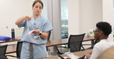 A female doctor in light blue scrubs in a classroom demonstrates suturing to high school students taking summer enrichment courses. Photo by K Pearlman Photography.