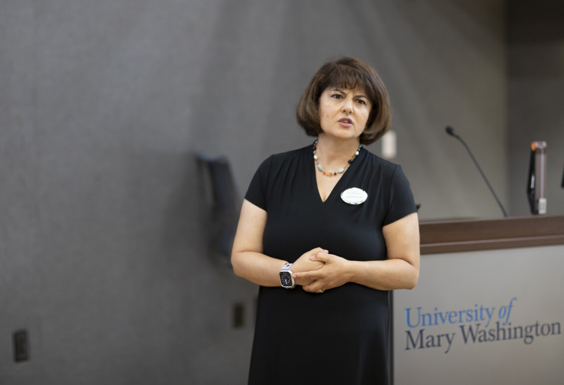 A woman in a black dress, wearing a name tag, speaks at the front of a classroom