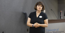 A woman in a black dress, wearing a name tag, speaks at the front of a classroom