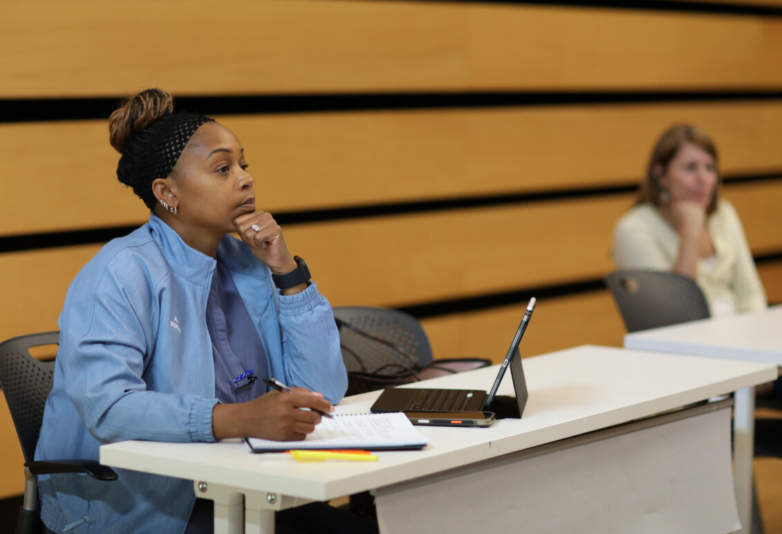 A woman sits at a desk with a pad of paper and a laptop.