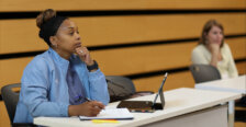 A woman sits at a desk with a pad of paper and a laptop.