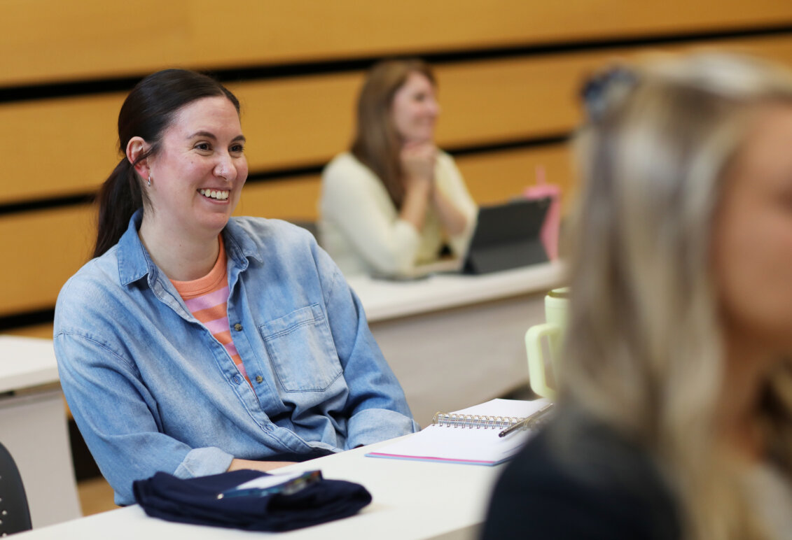 A woman wearing a denim shirt smiles as she sits in a classroom