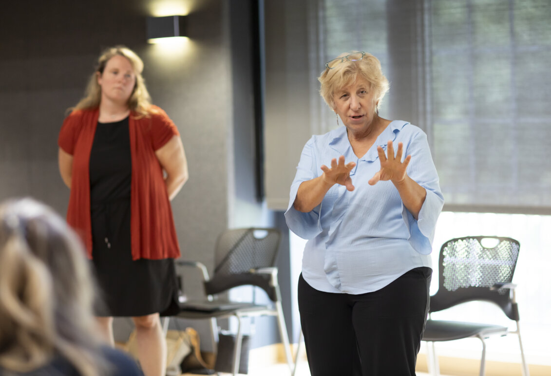 A woman in a light blue shirt uses her hands as she addresses a classroom, while another woman stands in the background