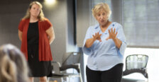 A woman in a light blue shirt uses her hands as she addresses a classroom, while another woman stands in the background