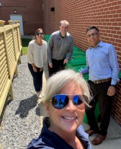 four people stand outdoors on a sunny day near the food composting bins next to a brick building on UMW's campus