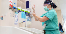 A female doctor in green scrubs with a face mask, clear goggles and hair covering stands in front of a sink scrubbing her hands to prepare to perform surgery. Photo by Parker Michels-Boyce.