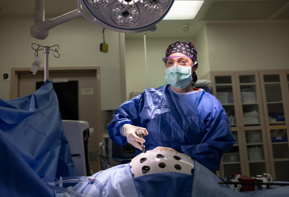 A female doctor in blue scrubs with a face mask, clear goggles and hair covering stands at a surgical table. Photo by Parker Michels-Boyce.