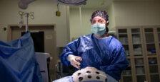 A female doctor in blue scrubs with a face mask, clear goggles and hair covering stands at a surgical table. Photo by Parker Michels-Boyce.