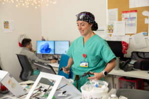 A female doctor in green scrubs with hair covering stands in a medical office room practicing with surgical instruments, a coworker in the background. Photo by Parker Michels-Boyce.