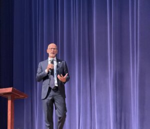 UMW President Paino in a blue suit and tie holds a microphone on stage in front of a blue curtain backdrop
