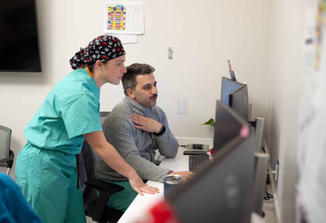 A woman in green scrubs and hair covering stands next to a male coworker in scrubs and a grey zip-ip jacket looking at a computer screen. Photo by Parker Michels-Boyce.