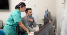 A woman in green scrubs and hair covering stands next to a male coworker in scrubs and a grey zip-ip jacket looking at a computer screen. Photo by Parker Michels-Boyce.