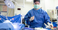 A female doctor in blue scrubs with a face mask, clear goggles and hair covering stands next to a surgical table with an instrument in her hand. Photo by Parker Michels-Boyce.