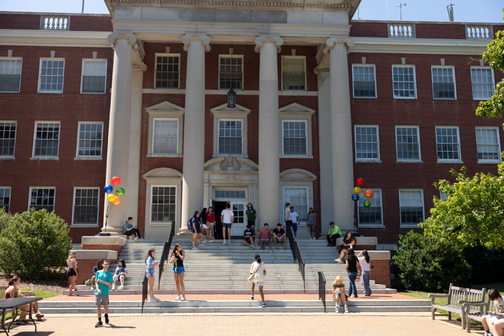 Several college students mingle outside a brick building with white columns