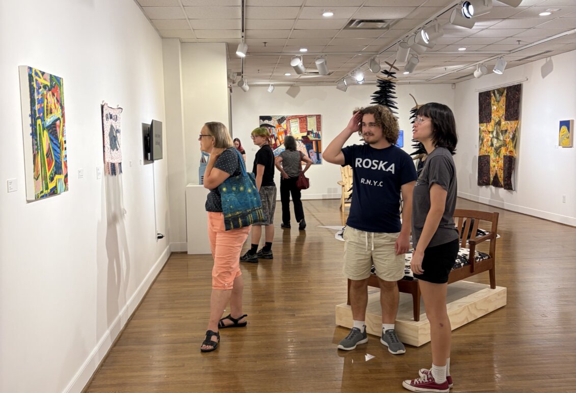 Visitors view artwork displayed along the walls of a gallery, with additional sculptures and installations visible throughout the room.