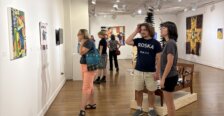 Visitors view artwork displayed along the walls of a gallery, with additional sculptures and installations visible throughout the room.